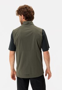 Olivgrüne Weste mit hohem Kragen, aus leichtem Stoff und einem schwarzen kurzärmeligen Hemd darunter. Auf der Rückseite befindet sich ein Logo in der Nähe des Saums.