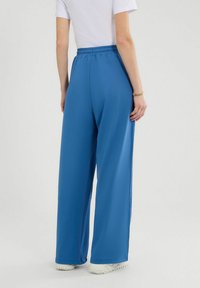 Lichtblauwe, loszittende broek met een elastische tailleband, gemaakt van een glad materiaal, gecombineerd met witte sneakers, gezien van achteren.