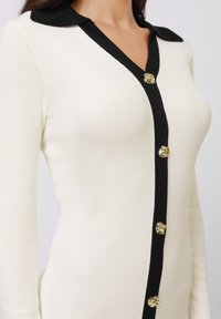 Hvit ribbestrikket cardigan med svart krage, med svart knappestolpe og gullknapper for lukking.