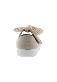 Zapato de lona beige con una suela de goma blanca, que presenta un lazo a rayas beige y blanco en una correa trasera. Textura suave y diseño sencillo.