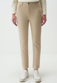 Pantalon beige slim en tissu mélangé coton avec cinq poches, passants de ceinture et fermeture à bouton. Porté avec des baskets blanches.