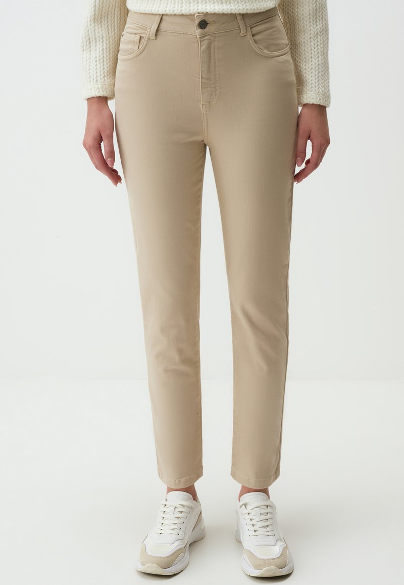 Pantalon beige slim en tissu mélangé coton avec cinq poches, passants de ceinture et fermeture à bouton. Porté avec des baskets blanches.