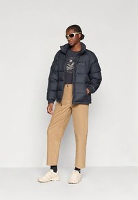 Fekete pufi dzseki magas gallérral, cipzáras zsebekkel, sötét grafikás póló fölött viselve, bézs nadrággal és világos sportcipőkkel párosítva.