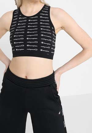Kvinde iført sort crop top med gentagne hvide "Champion" logoer og sorte joggingbukser med Champion logo på hoften.
