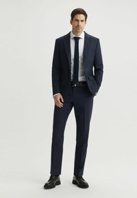 Granatowy, dopasowany garnitur z klapami w szpic, dopasowaną krawatem i białą koszulą. Wyróżniające się fakturowane akcenty oraz dopasowane spodnie. Czarne buty.
