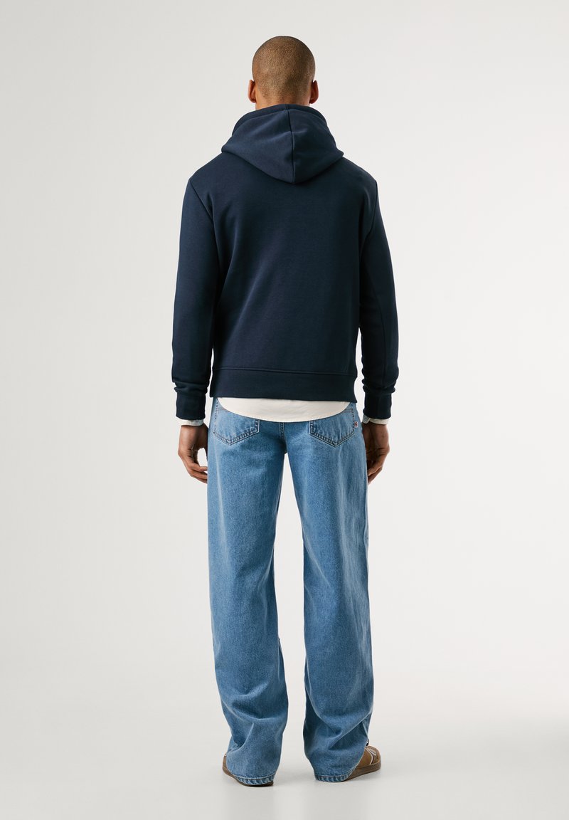 Marineblauwe hoodie met een grote capuchon, zachte textuur en geribbelde manchetten, gecombineerd met lichtblauwe wijde jeans en bruine schoenen.