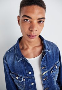 Denim jacket in medium blue with metal buttons, chest pockets, and a classic collar, layered over a white tank top. Smooth texture, casual style.