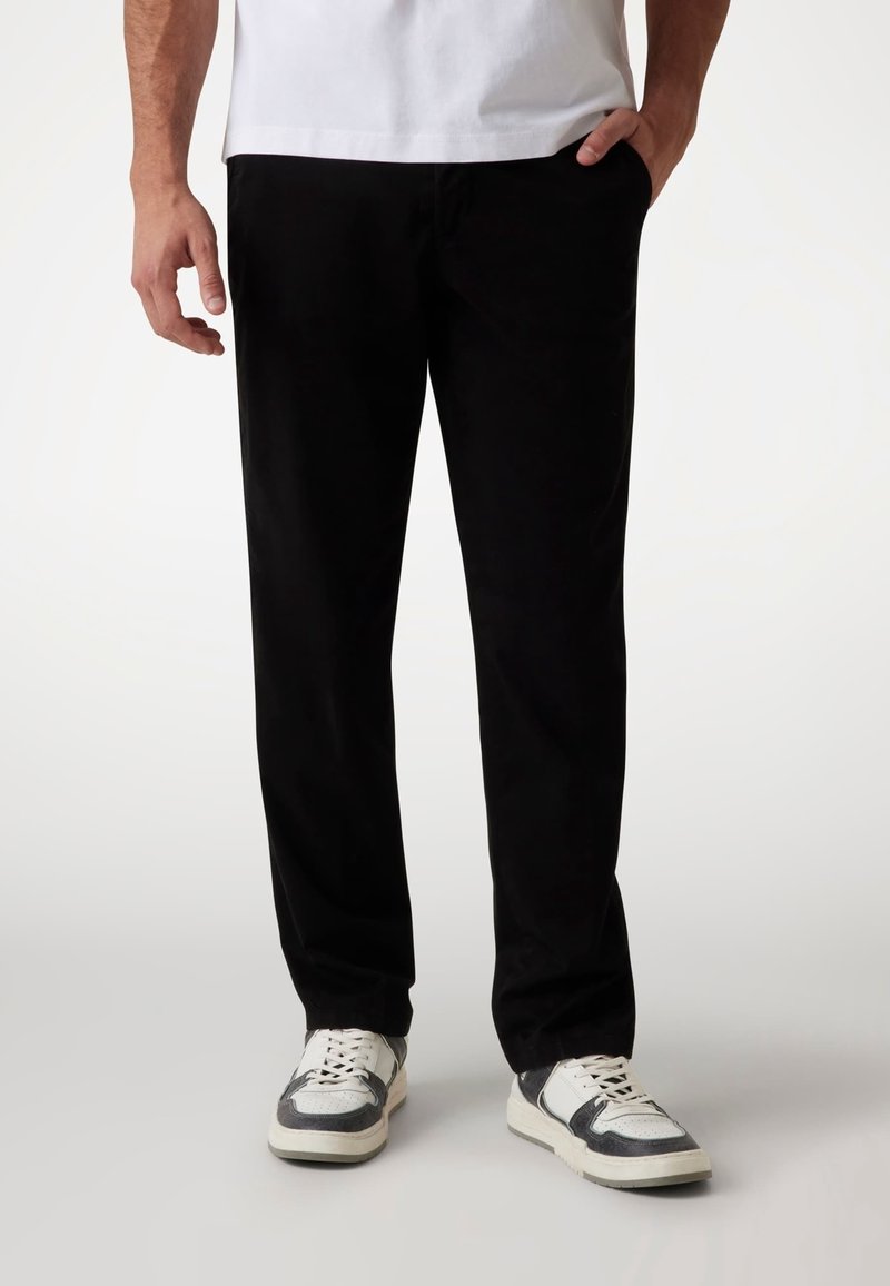 Pantalon en coton noir à coupe droite, doté de poches latérales, porté avec des baskets blanches et grises, associé à un t-shirt blanc uni.