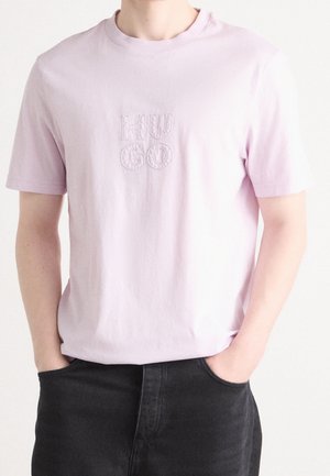 Person trägt ein hellrosa kurzärmeliges T-Shirt mit erhabenen "HUGO" Buchstaben in der Mitte der Brust, Hände in den Taschen einer schwarzen Hose.