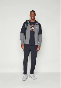 Sportsoutfit bestående af en grå og sort lynlåsjakke, grafisk sort t-shirt og mørke bukser med reflekterende detaljer. Hvide sneakers fuldender looket.