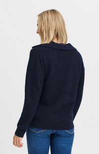 Sötétkék textúrált pulóver, laza szabással, hosszú ujjal és széles gallérral, fitted kék farmernadrággal párosítva.