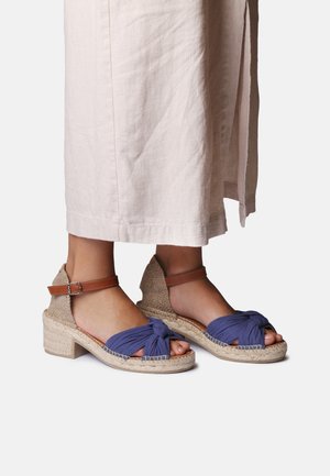 Pieds portant des sandales espadrilles en tissu bleu à nouer sur le devant, avec des brides marron à la cheville et des talons tissés bas, assorties à une longue jupe beige.