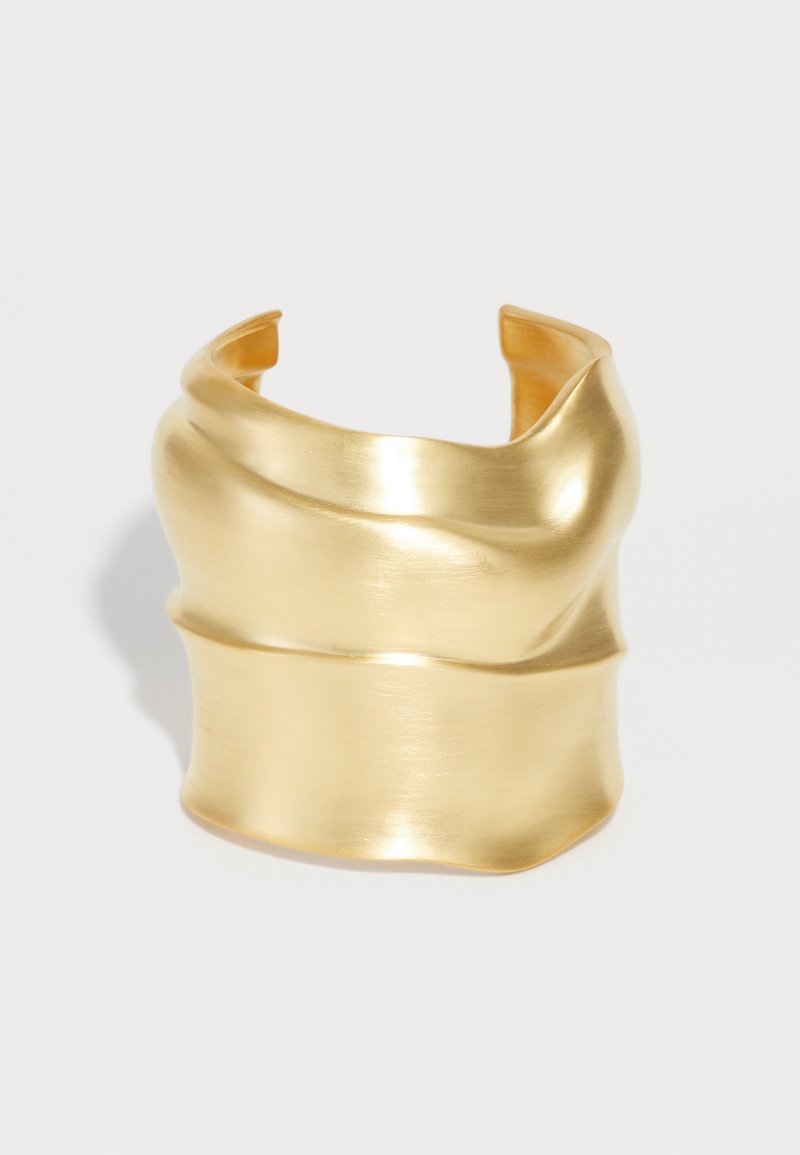 Βραχιόλι μανσέτας σε χρυσό χρώμα με κυματιστό, γλυπτικό σχέδιο. Λείο φινίρισμα από βουρτσισμένο μέταλλο, ανοιχτή πλάτη και οργανικό σχήμα χωρίς ορατούς συνδετήρες.