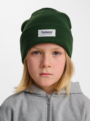 Kind met lang blond haar dat een donkergroene muts draagt met het label "hummel sportswear" en een grijze zip-up hoodie, neutrale uitdrukking.