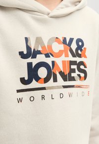 Bež kapucar s izrazito večbarvno grafiko z napisom "JACK & JONES WORLDWIDE" v krepki pisavi in različnih vzorcih.