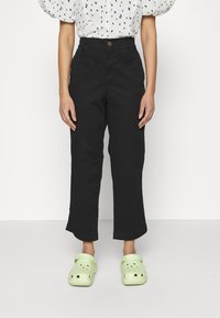 Pantaloni neri a gamba larga con vita alta e chiusura a un bottone. Indossati con zoccoli verdi chiari e una camicia bianca a pois a forma di cuore.