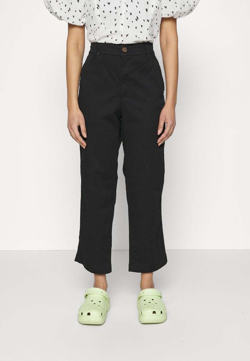 Pantaloni neri a gamba larga con vita alta e chiusura a un bottone. Indossati con zoccoli verdi chiari e una camicia bianca a pois a forma di cuore.