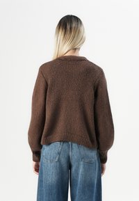 Person med langt blondt hår iført en brun strikkegenser og blå denimjeans, vendt bort fra betrakteren mot en hvit bakgrunn.