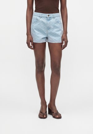 Kvinne iført lyseblå polkadott denimshorts, brun ermeløs topp og brune kilehælssandaler, stående mot en ensfarget bakgrunn.