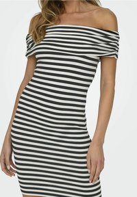 Nainen pukeutuneena vartalonmyötäiseen olkapäät paljastavaan mekkoon, jossa on vaakasuorat mustavalkoiset raidat, yksinkertaisen taustan edessä.