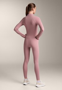 Rosa träningsoutfit bestående av en åtsittande långärmad tröja och tights, tillverkad av elastisk tyg, tillsammans med vita sneakers.