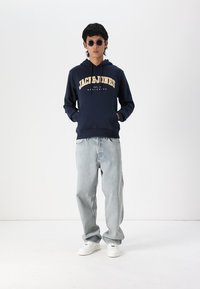 Marine hoodie met het "JACK & JONES" logo in geel, gecombineerd met lichtblauwe jeans en witte sneakers. Model draagt zwarte zonnebril.