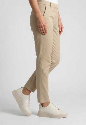 Person iført beige skræddersyede bukser og hvide perforerede sneakers, stående på en lysegrå baggrund.