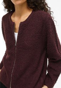 Burgundska zip-up jakna z teksturirano, puhasto tkanino in rebrastim ovratnikom. Ima dolge rokave in srebrn zadrgo.