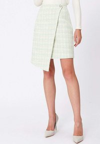 Femme portant une jupe en tweed asymétrique vert pâle avec un bouton frontal, associée à des talons hauts blancs et un haut blanc à manches longues.