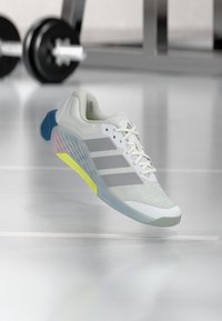 Λευκό αθλητικό παπούτσι με επάνω μέρος από πλέγμα, γκρι ρίγες και πολύχρωμες λεπτομέρειες σε μπλε και κίτρινο. Διαθέτει υφή στη σόλα και ελαφρύ σχεδιασμό.