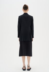 Blazer negro ajustado de textura suave, con una sola abertura en la parte posterior y puños abotonados, combinado con una falda negra hasta la rodilla y sandalias planas.