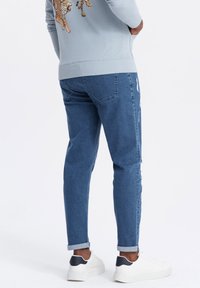 Persona vestida con sudadera azul claro con diseño de tigre, jeans azules remangados y zapatillas blancas con detalle negro en el talón, de pie de lado.