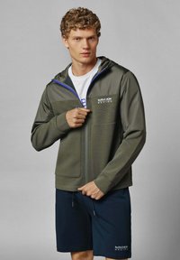 Maslinasto zelena jakna sa kapuljačom na zip, s teksturiranom površinom i kontrastnim plavim detaljima, u kombinaciji s tamnim šortsevima. Prikazan Red Bull Racing logo.