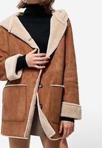 Chaqueta de ante marrón con una capucha forrada de suave tono beige, grandes bolsillos y dos botones. Las mangas también están forradas con pelo sintético beige.