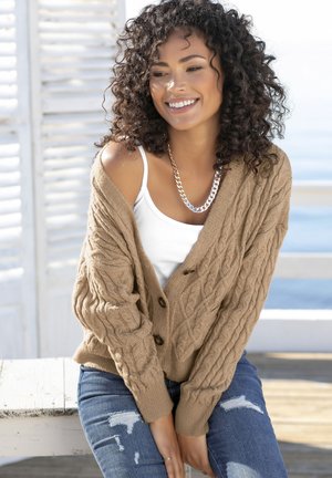 Brauner Strickcardigan mit zwei Knöpfen, über einem weißen Tanktop getragen. Kombiniert mit ausgewaschenen blauen Jeans. Natürlicher Hintergrund.