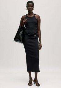 Schwarzes, geripptes Tanktop-Kleid mit verstellbaren Trägern, einem runden Akzent an der Taille, kombiniert mit einer glänzenden schwarzen Tote-Tasche und sandalenartigen Schuhen.