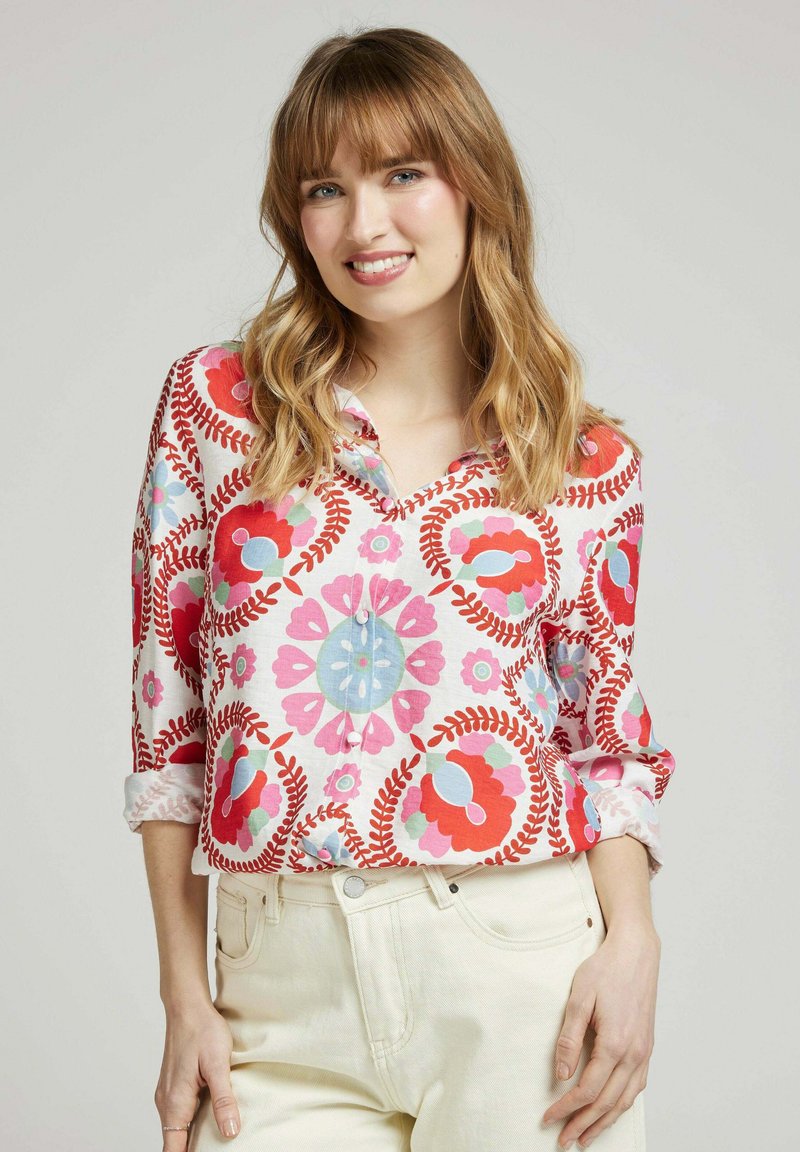 Chemise à boutons avec un motif floral coloré en rouge, rose et bleu sur un tissu blanc ; possède des manches retroussées et un col bandé.