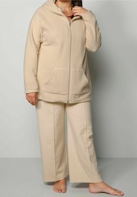 Sudadera de forro polar beige con cremallera y dos bolsillos delanteros, con una capucha forrada, combinada con pantalones anchos beige que muestran una textura suave.