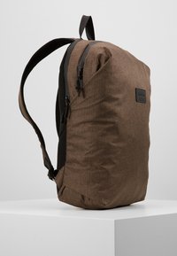 Zaino in tessuto marrone dal design elegante, caratterizzato da materiale testurizzato, una tasca laterale in rete e due cerniere nere lungo la parte superiore.