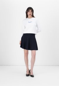 Felpa bianca a maniche lunghe con testo blu e gonna plissettata blu navy, abbinata a tacchi a punta neri, su uno sfondo semplice.