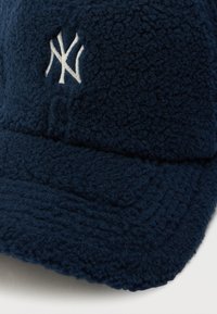 Tumši zila flīsa cepure ar paaugstinātu baltu logotipu. Mīksta, teksturēta virsma ar noapaļotu mala un kontūrveida dizainu, kas nodrošina labu piegulšanu.