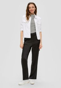 Witte denimjack over een zwart-wit gestreepte top, gecombineerd met zwarte wijde cargobroek en witte sneakers, tegen een grijze achtergrond.