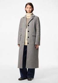 Mladá žena v dlouhém šedém vlněném kabátu, krémovém roláku, tmavých širokých kalhotách a černých botách stojící na bílém pozadí.