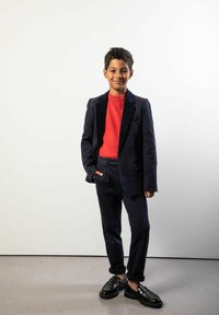 Granatowy garnitur z czarnymi aksamitnymi klapami, pod spodem czerwony sweter, ciemne spodnie i czarne mokasyny. Młody mężczyzna pozuje z rękami w kieszeniach.