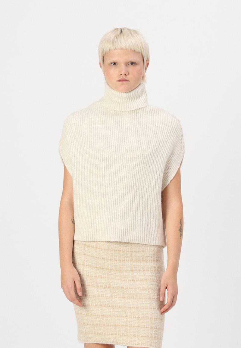 Kremna ribana pletena turtleneck pulover s kratkimi rokavi. Model ga kombinira z teksturirano bež krilom. Mehka, udobna tkanina.