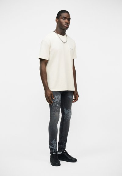 Άνδρας που φοράει εκρού μπλουζάκι, σκούρο τζιν με σχέδια και στενή γραμμή, μαύρα αθλητικά παπούτσια και ασημένιο κολιέ αλυσίδα, στέκεται μπροστά σε λευκό φόντο.