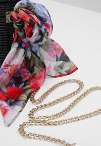 Sciarpa in seta floreale con un mix di fiori rossi, rosa e viola su uno sfondo nero, legata a una borsa nera intrecciata con una tracolla dorata.