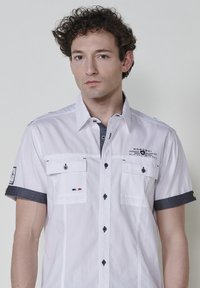 Camisa blanca de manga corta con cuello y puños oscuros, que presenta dos bolsillos en el pecho, detalles de botones y acentos bordados.