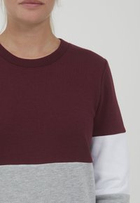 Bordo i siva pamučna dukserica s okruglim izrezom. Sadrži odeljke u blokovima boja i glatku teksturu. Kratki rukavi, minimalni dizajn.