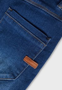 Σκούρα μπλε τζιν παντελόνι με εμφανή τσέπη και αντίθετη πορτοκαλί ραφή, καθώς και μικρή καφέ δερμάτινη ετικέτα.